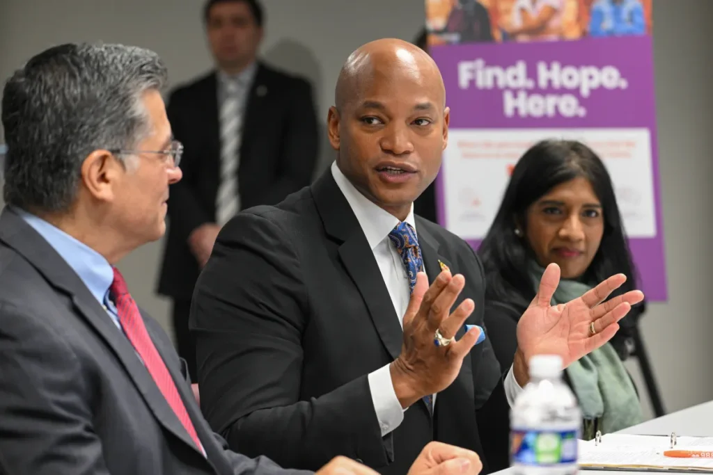 A man with gray hair and a woman with dark hair are seated at a table and listening to Maryland Governor Wes Moore, who is sitting between them.